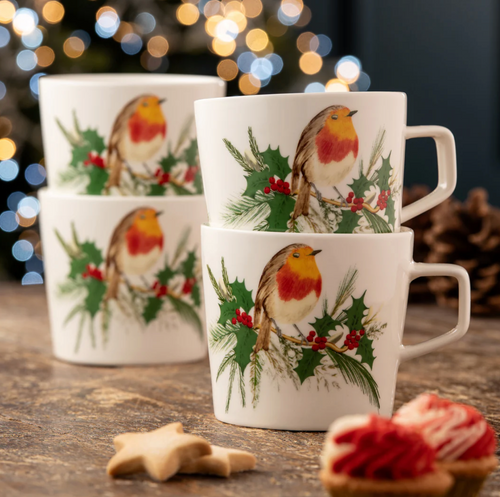 Set of three mugs with robin and holly design on a wooden surface with blurred lights in the background.