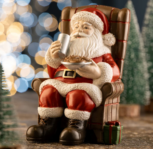 Santa Claus figurine sitting in a chair with a blurred Christmas tree and lights in the background
