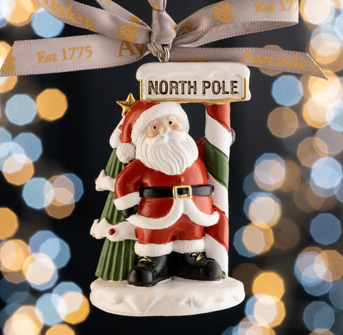Decorative Christmas ornament of Santa Claus holding a 'North Pole' sign with a blurred lights background.